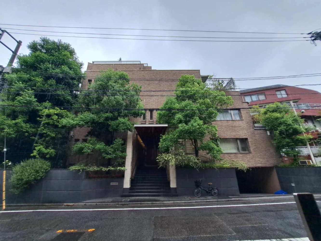 - 都会の中の緑が豊かな三階建ての brick風 exterior、正面の階段と入口、周囲に生い茂る木々と住宅街の風景