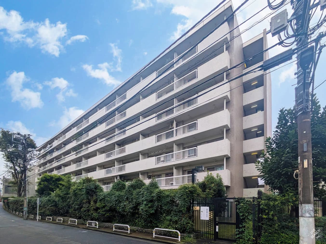 - 都市部の長方形の白い exterior を持つマンション。外観は水平のバルコニーが連続し、手前には緑豊かな植栽と黒の門がある。