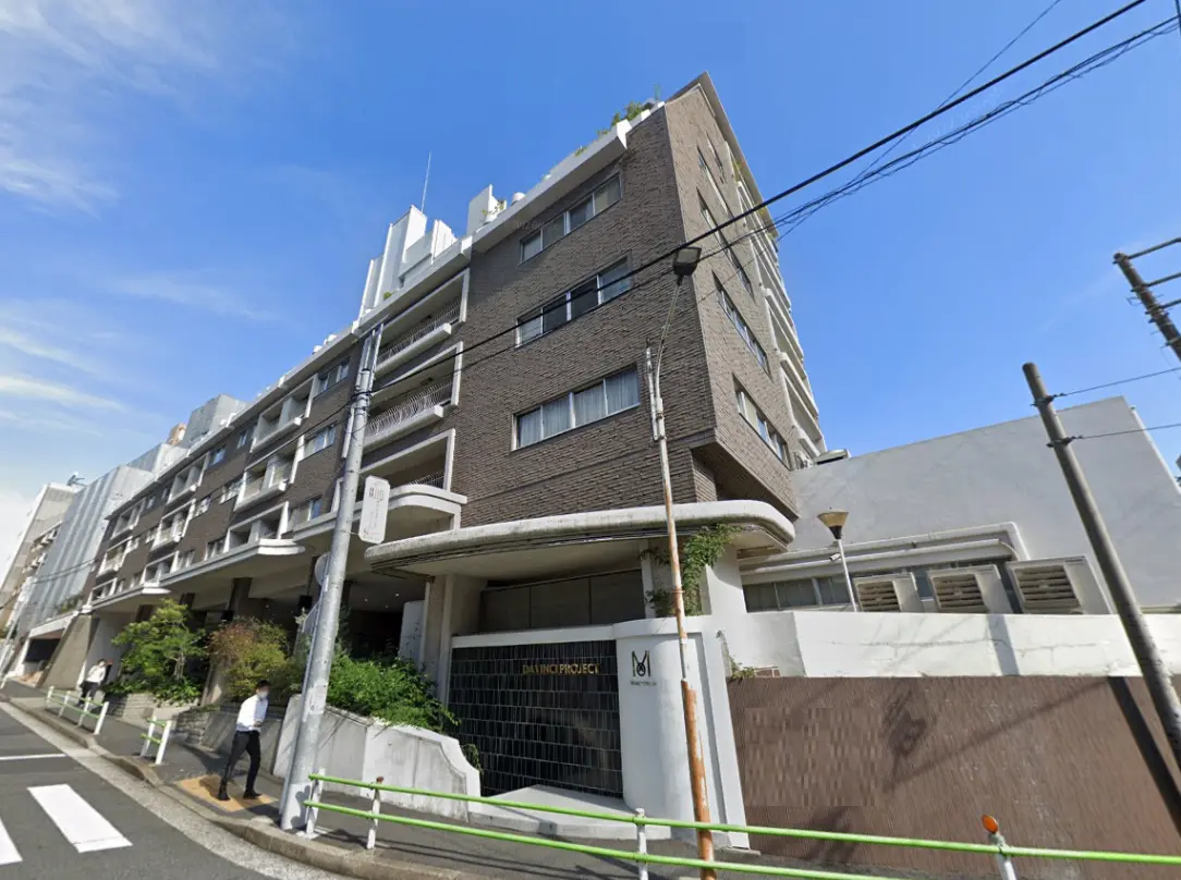- モダンな外観のマンション、茶色のタイル張りの壁、周囲に植栽と歩行者、青空の下での風景