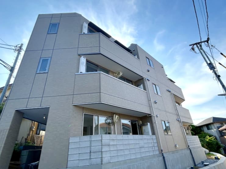 - 三階建ての現代的なマンション exterior、淡いベージュ色の外壁と連結したバルコニー、青空と周囲の住宅が背景に広がる風景