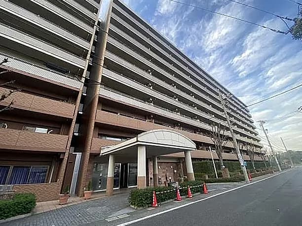 - 高さのあるブラウンの大型マンション exterior、エントランスへ向かう道沿い、白いアーチ型屋根の入口と周囲のコーン、都市部の通りの景観