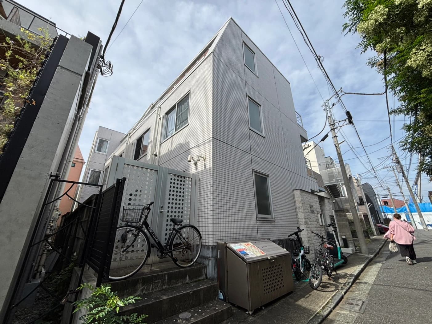 - 白いモザイクタイルの三階建て集合住宅が並ぶ路地風景、建物横に自転車とゴミ箱、電線が張り巡らす空と青空が広がる街並み