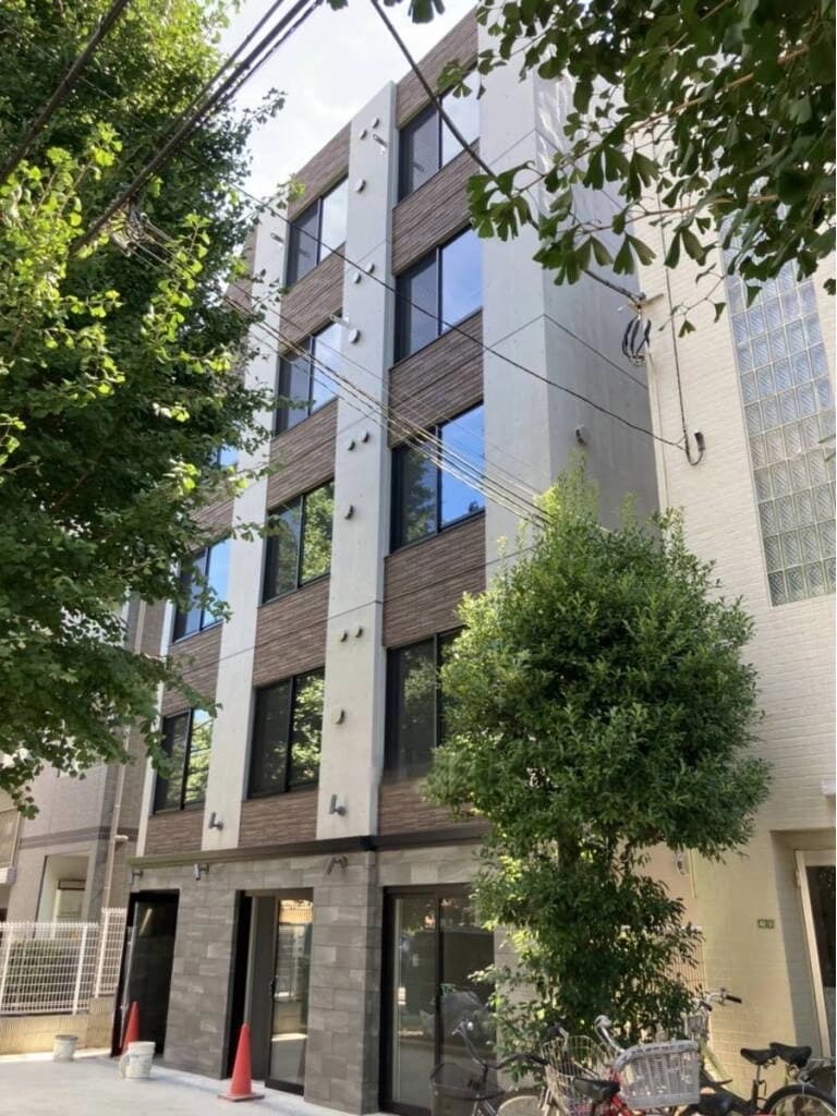 - 都市型の四階建てマンション外観、縦長の窓と白と茶系の外壁、周囲に木々と自転車が置かれた風景