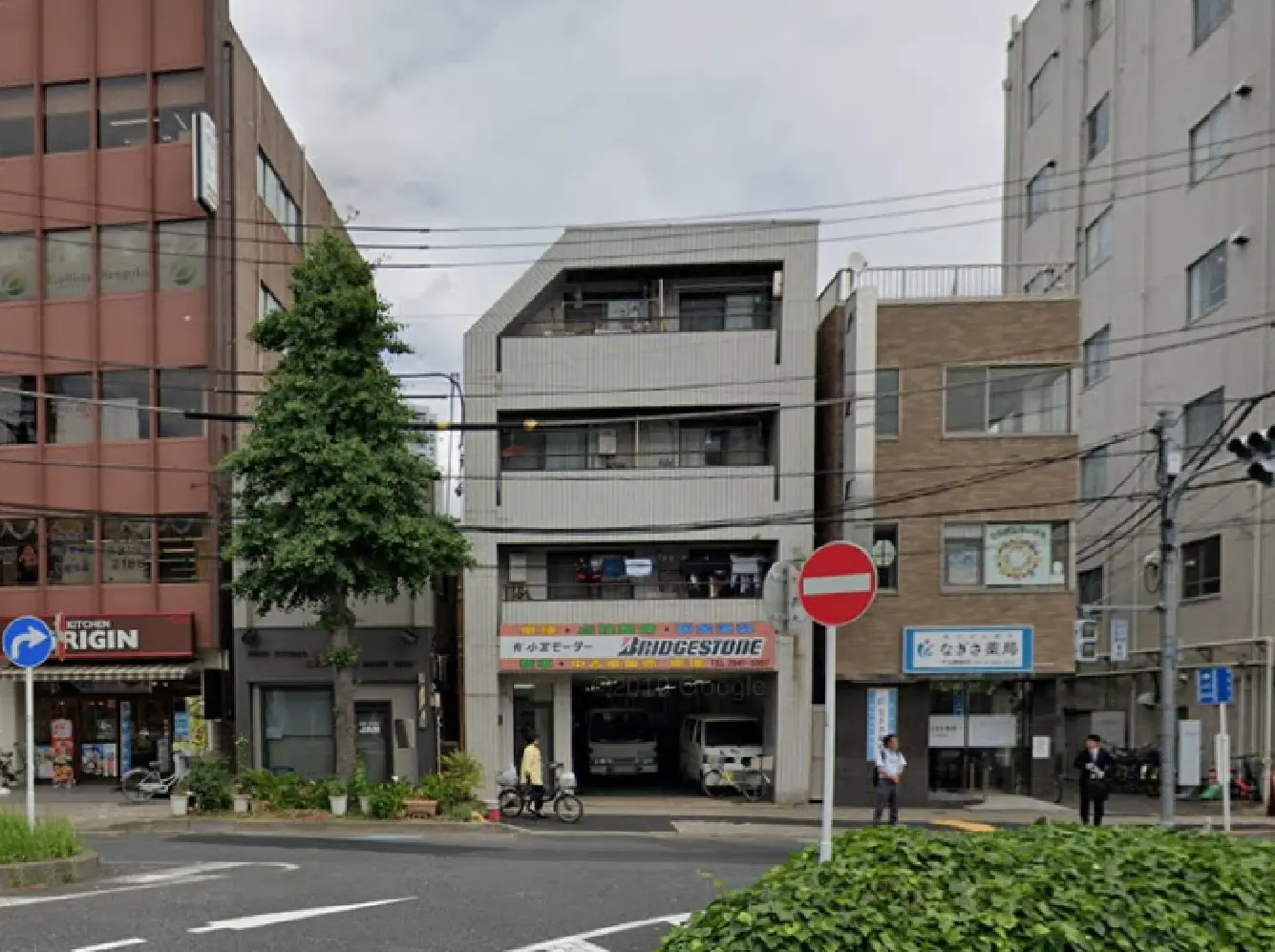 - 都市部の商業施設が並ぶ交差点、ブリヂストンの看板が掲げられた建物と緑の木々、周囲に捨てられた車両が見える風景