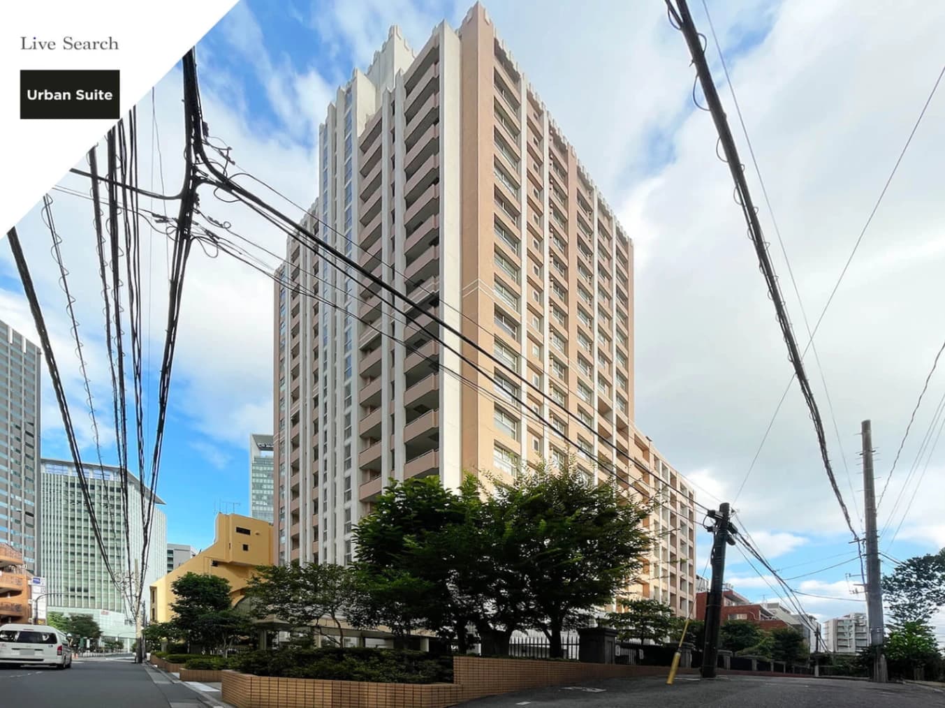 - 都市の高層マンションと周囲の電線、青空と路地風景、緑の植栽が特徴的な集合住宅外観の写真
