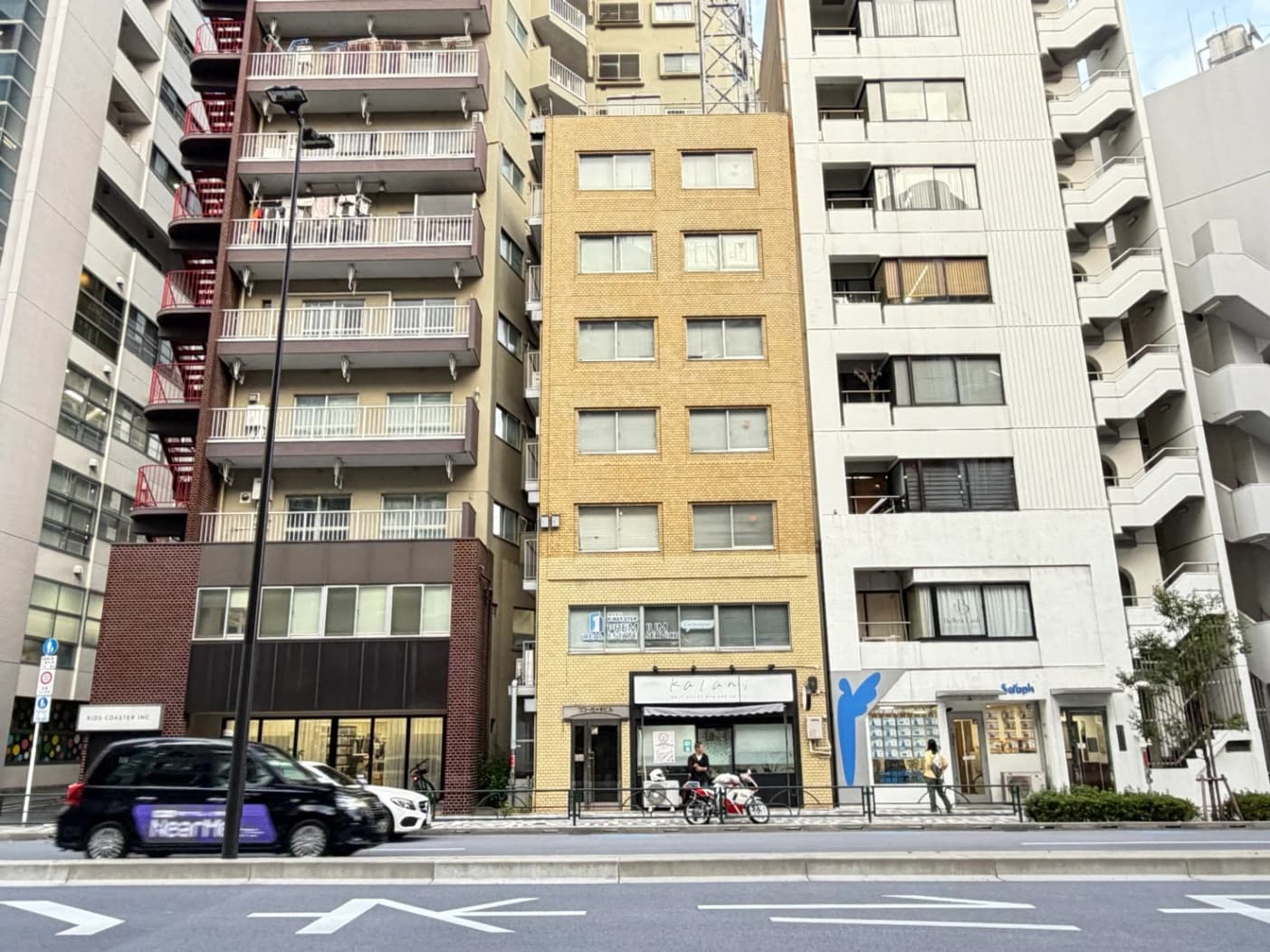 - 都市の通りに立つ中高層の建物群、中心にベージュ色のレンガ造りの4階建てビルがあり、両サイドに白とグレーの現代的な高層マンションが並ぶ光景
- 路面電車通りの横断歩道前、窓が並ぶビル群と店舗が1階に連なる街角風景、駐車中の車と自転車、歩道には看板や小さな木々が点在
- ベージュのレンガ造りビルの正面、窓が均等に配置された4階建て、1階には店舗の看板とガラス入口、周囲は高層マンションと道路、交通標識が見える
- 通り沿いの建物群、左に茶色の外観のビル、中央にベージュのレンガビル、右に白系の建物が続く風景、前景には車と自転車が走行中