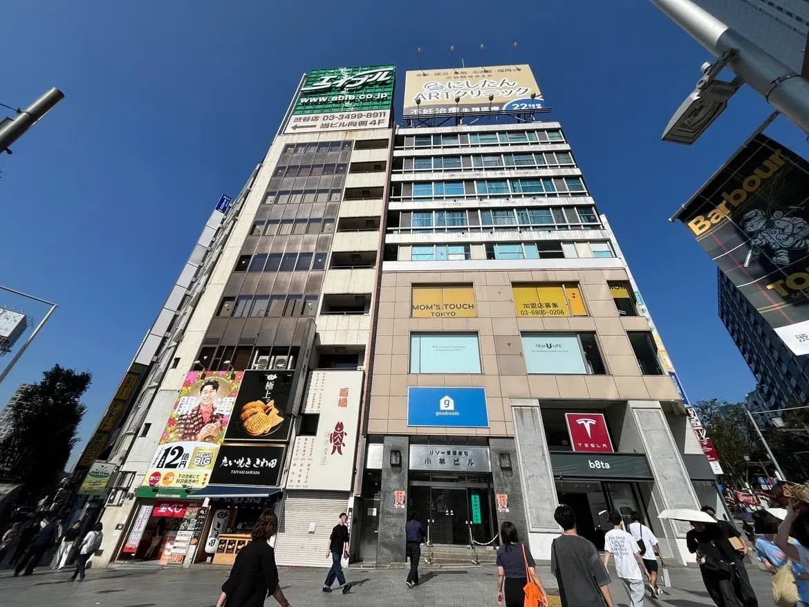 - 渋谷の繁華街にある多階建てのビル、様々なショップの看板が並ぶ、青空の下を行き交う人々の風景