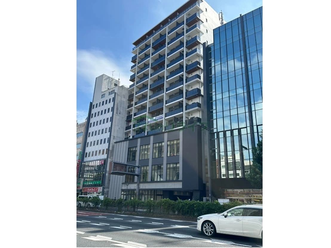 - 都心の高層オフィスビルとテナント店舗が並ぶ通り沿いの風景、ガラス張りの高層ビルと白い建物、路上を走る白い車と緑の植栽
