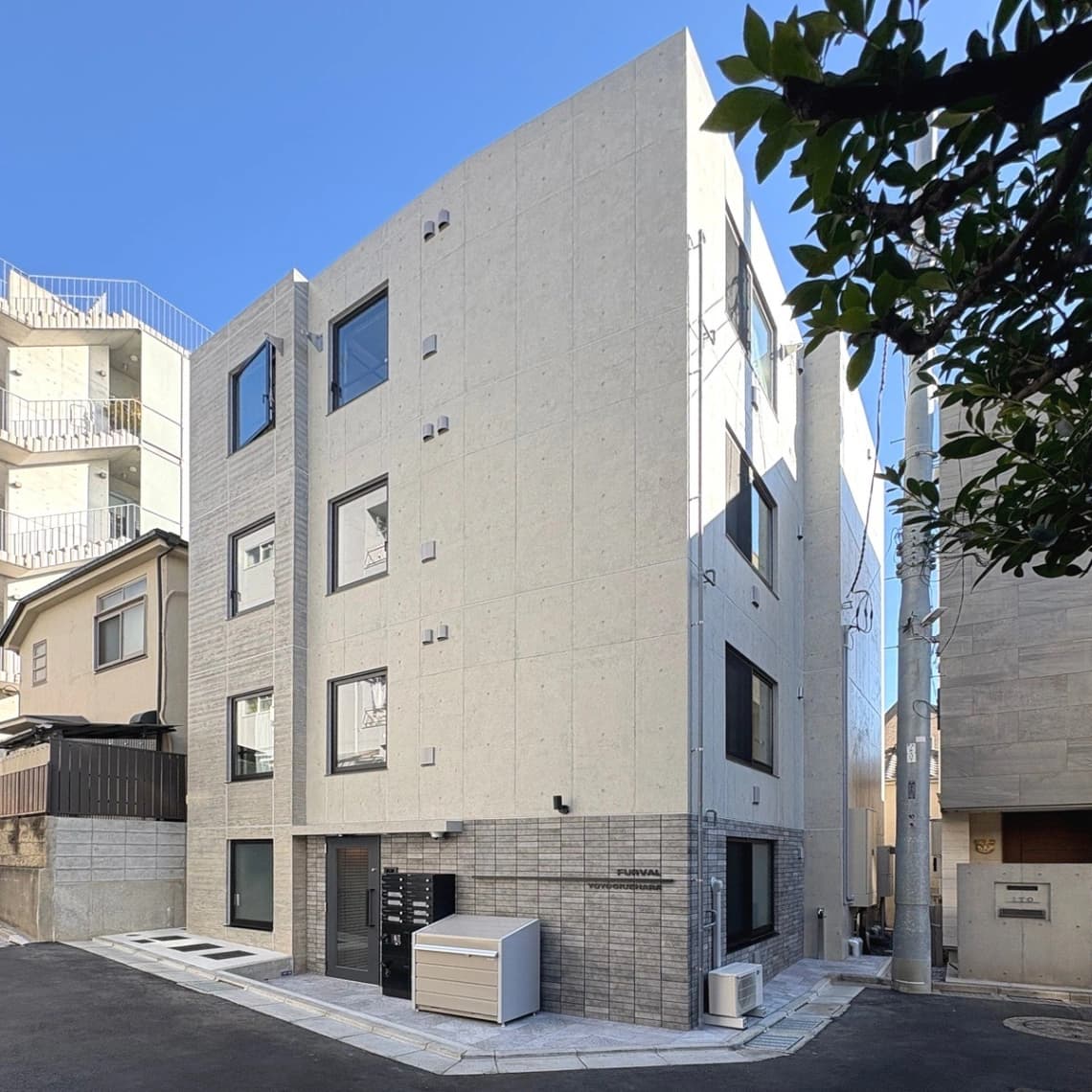 - 都市の四角い façade を持つモダンな集合住宅。石張りの低層部と窓が配置された白い外観、狭い路地と隣接建物、青空と木々の影が映える写真