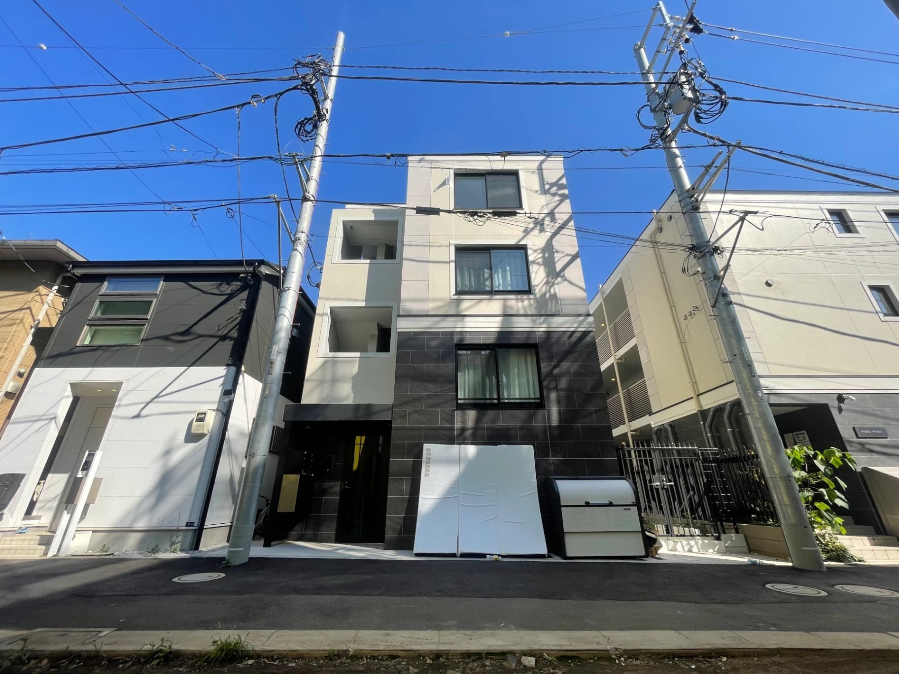 - 現代的な白と黒の三階建てマンション群、前方に電信柱と多くの電線が crossed、青空と影が映える都市風景