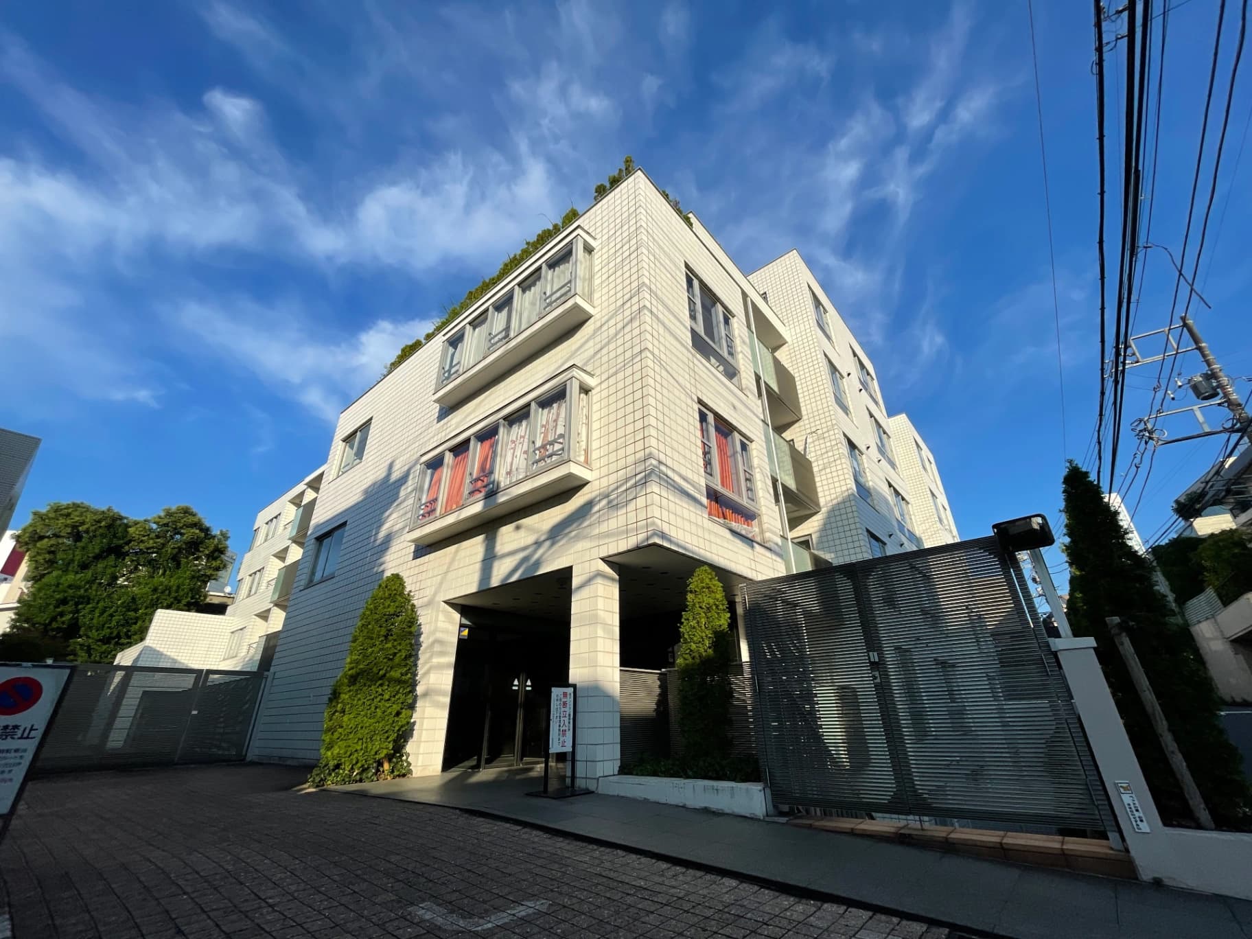 - 白いタイル張りの高層マンション外観、角度をつけたファサードと大型窓、青空と電線が背景に映える都市景観