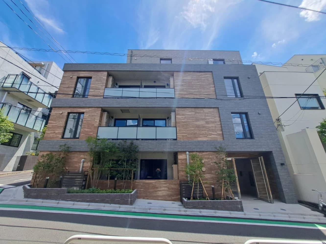 現代的な3階建てマンション外観、ブラウンの石目調とグレーのタイル張りで窓とバルコニーが配置され、正面には緑の植栽とエントランスドアがあり周囲には都市的な街路が広がる外観写真