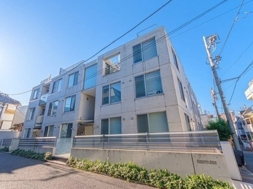 コンクリート打ちっぱなしの三階建て新築風のマンション外観、各階に大きな窓とベランダがあり、手すり付きの安定した外周フェンスと敷地内の低木が整備された都市型アパートメント