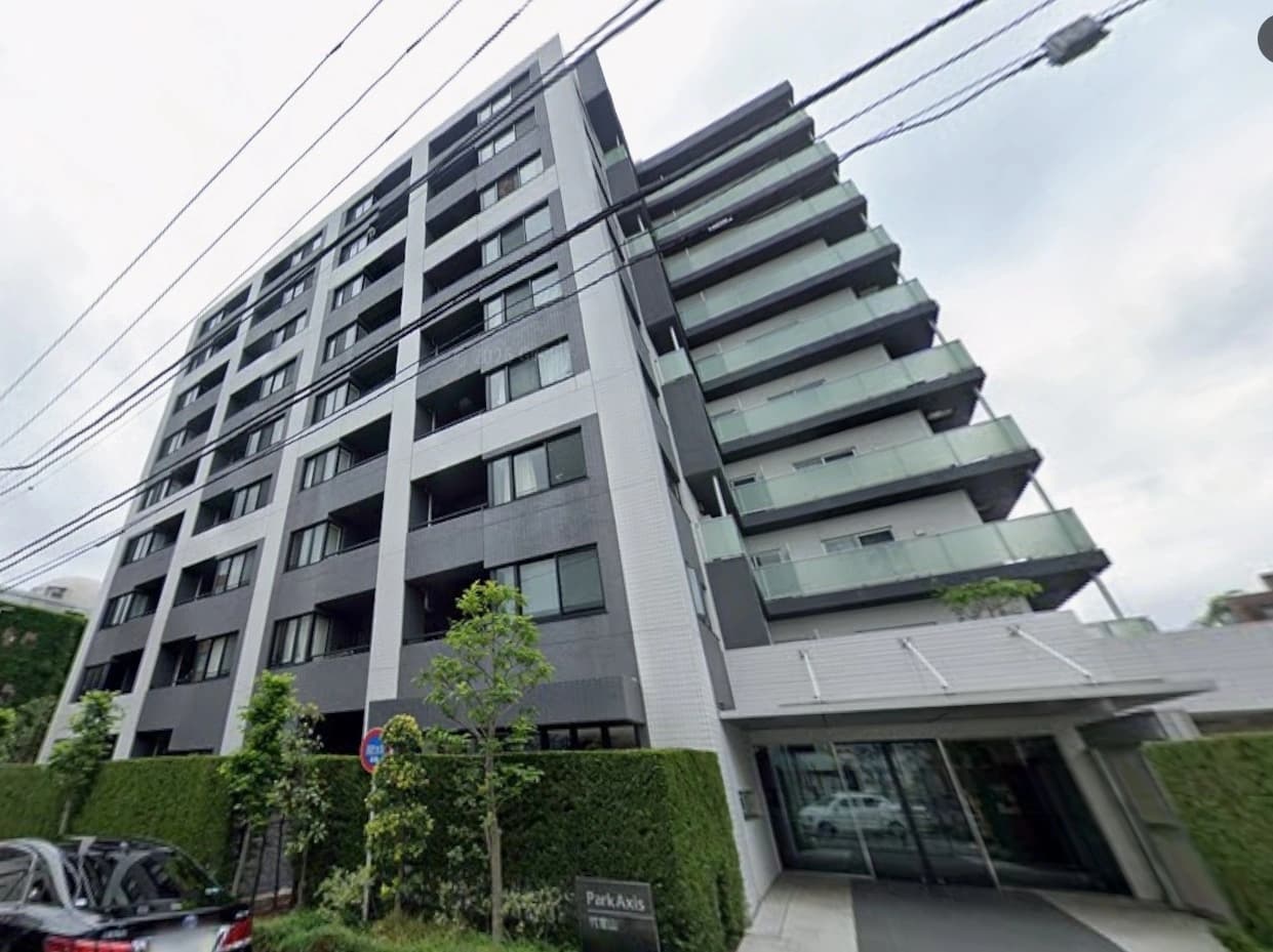 - 都心部の高層マンション外観、ガラス張りのバルコニーと白黒の外装デザイン、入り口前の緑の植栽と駐車スペースが映る都市景観