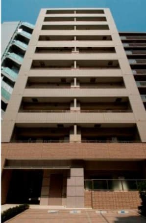 - 高さのある鉄筋コンクリート造のマンション外観、幅広い階層と縦の連続窓、新緑の装飾とエントランス前のタイル床の建物写真