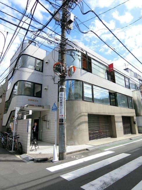 - 都市の角地にある三階建ての白いタイル張りビルと交差点、電線が風景の上部を覆い、入口付近に自転車と歩行者、大きな窓ガラスと車線のある通りの外観