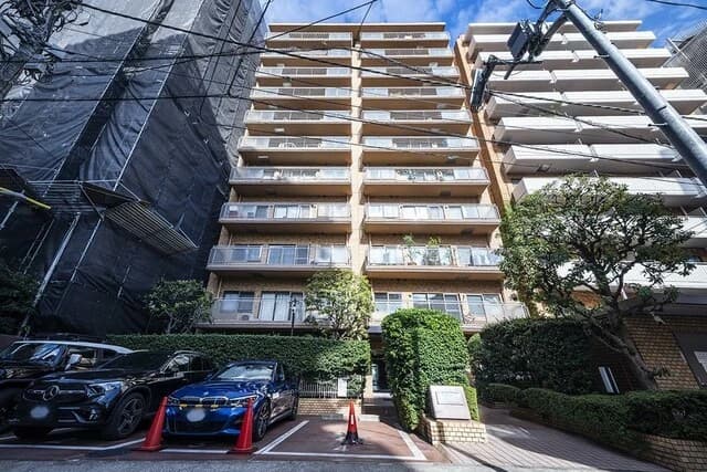- 高層マンションの外観、手前に車両とコーンが並ぶ駐車場、左右に樹木と低い生垣があり、透明ガラスのバルコニーが連なる都市型住宅街の光景