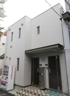 - 白いモダンな2階建て住宅 exterior、入口横に自動販売機と郵便受け、整然としたタイル張りの前庭とやや angled のファサード