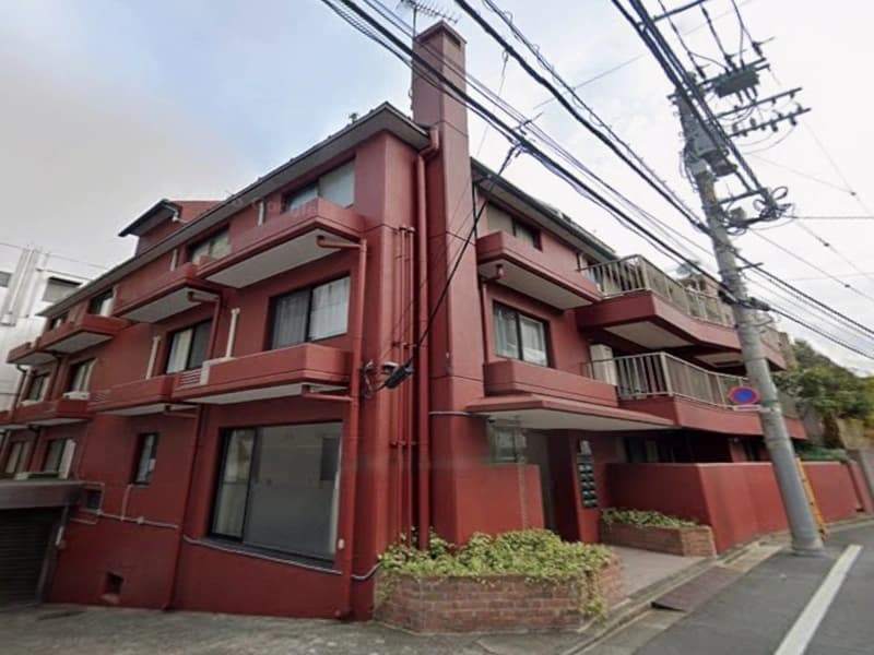 - 茶系の三階建てマンション exterior、赤茶色の外壁とバルコニー、路地沿いの小さな植栽と入口付近のレンガ花壇