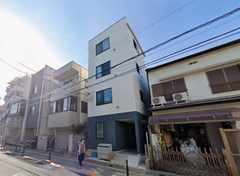 専用階段のある3階建ての新築風の白と濃いネイビーブルーの外観が特徴的な三階建て住宅が路地沿いに立ち並ぶ街並みの風景