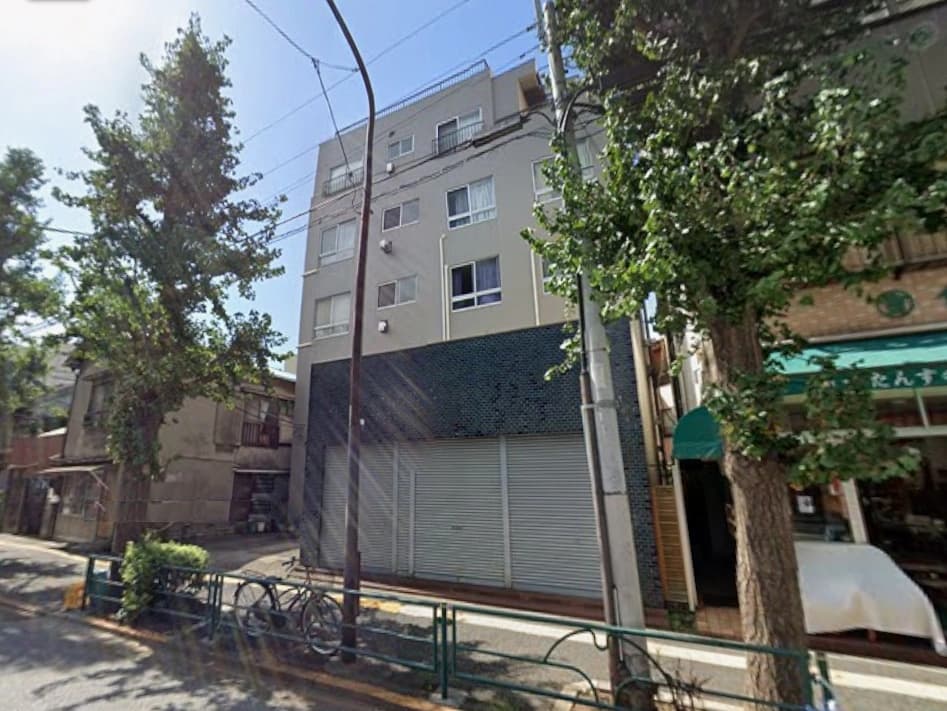 - 都心の四階建てビル外観、路面にはシャッター商店と緑の木々、通り沿いの風景と自転車が見える街並み
