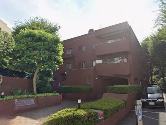 - 赤茶色の三階建てマンション exterior、緑豊かな樹木と低い生垣に囲まれ、車が駐車場脇に停車している風景