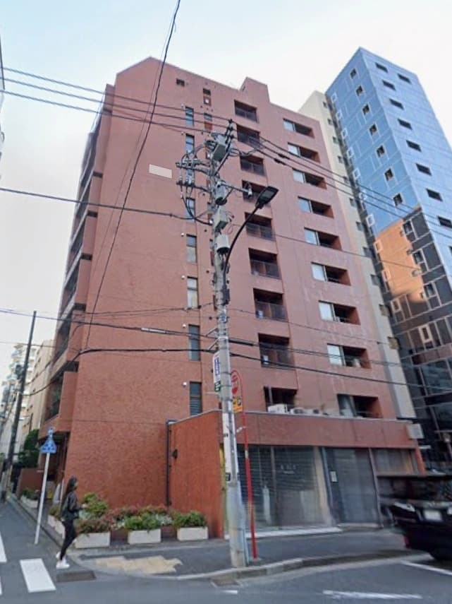 - 都市部の角地に立つ複数階建ての赤茶色のマンション。低層部にはガラス窓の商業スペースがあり、周囲には電柱と配線が交差する光景