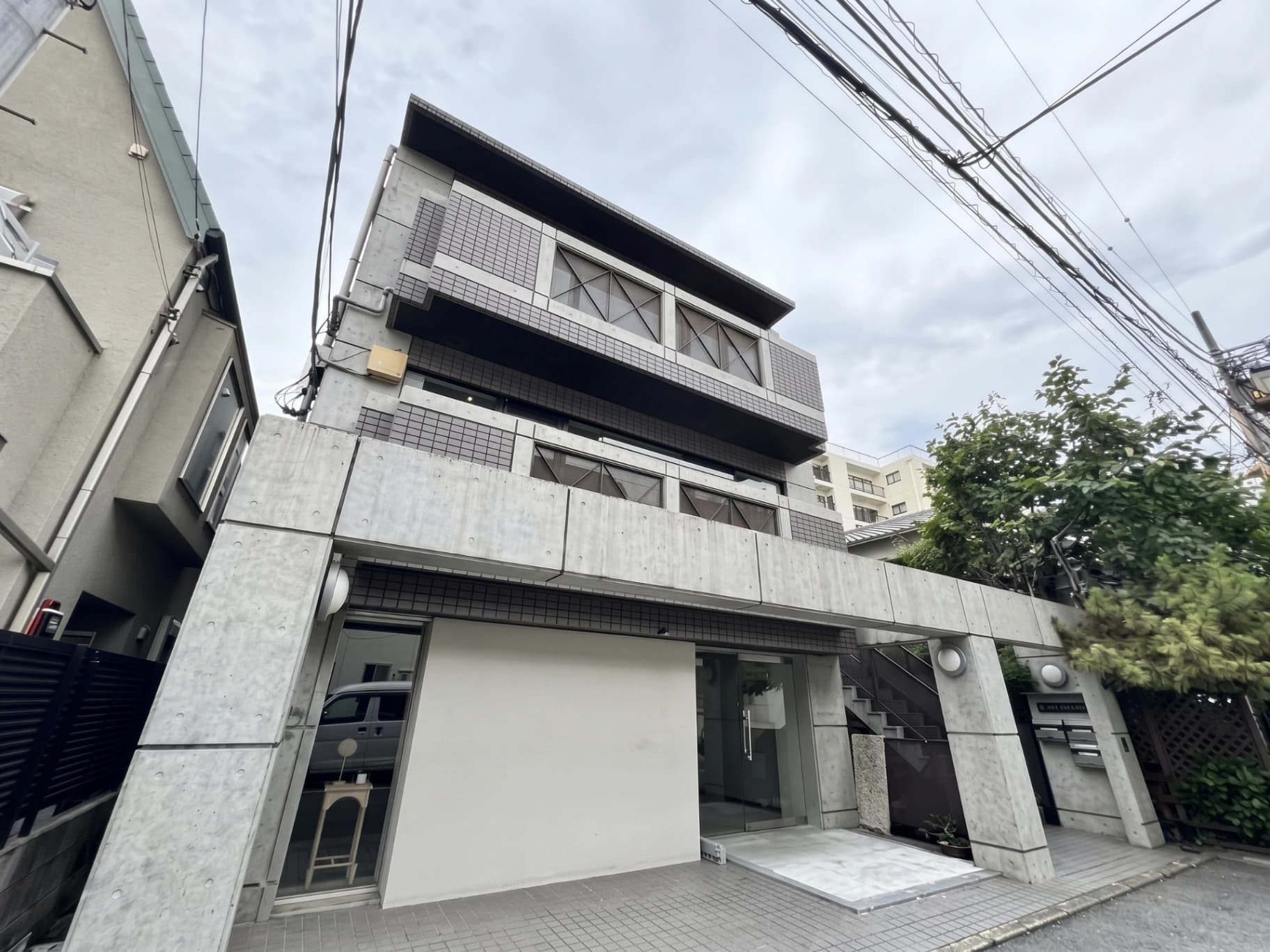 - コンクリートとガラスを多用した現代的な3階建てマンションの外観、ファサードにはグリッド状のタイルと大きな窓、前庭には植栽と駐車スペース、上空には電線が走る街並みの風景