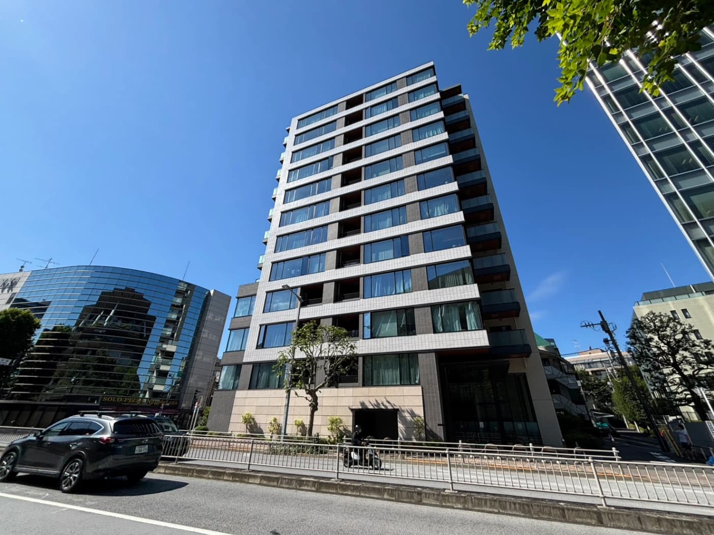 - 都市の繁華街に立つ白と灰色の高層マンション、ガラス窓が整然と並ぶ現代的な外観と晴天の青空
