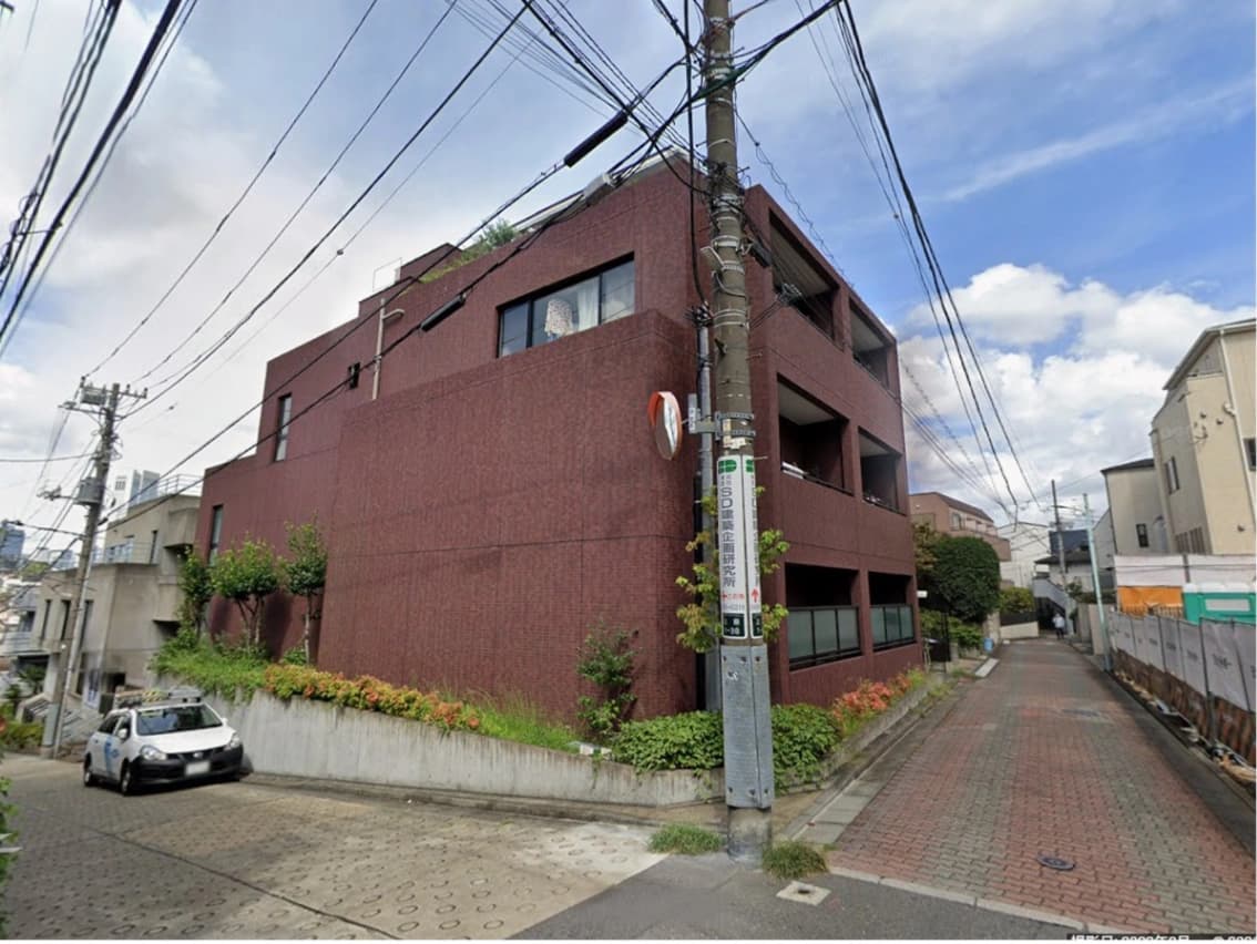 - 赤茶色の角地に建つモダンな3階建てマンション風建築と周囲の住宅街、電線が空へ伸びる街路と石畳の道、緑の植栽と青空の風景