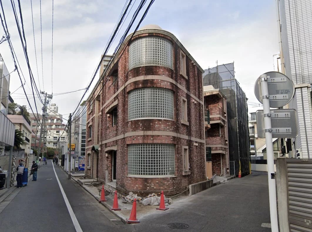 赤レンガ造りの円形が特徴の3階建てビルが路地沿いに建ち、周囲には電線が多数張り巡らされ、工事用コーンと建設資材が道路端に置かれた都市景観