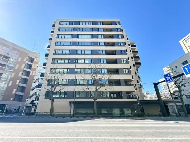 - 高層の現代的なマンション外観、複数階にわたる窓とバルコニーが並ぶ都市景観、手前に樹木と広い sidewalk、青空の下に伸びる建物