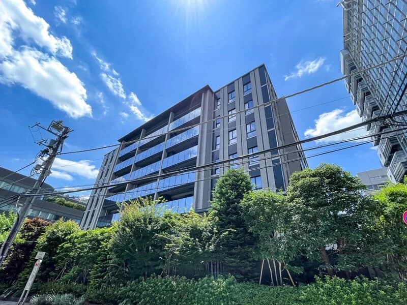 - 都心の高層オフィスビル外観、ガラス張りの窓とモダンなデザイン、周囲に緑豊かな植栽と電線が見える青空の写真