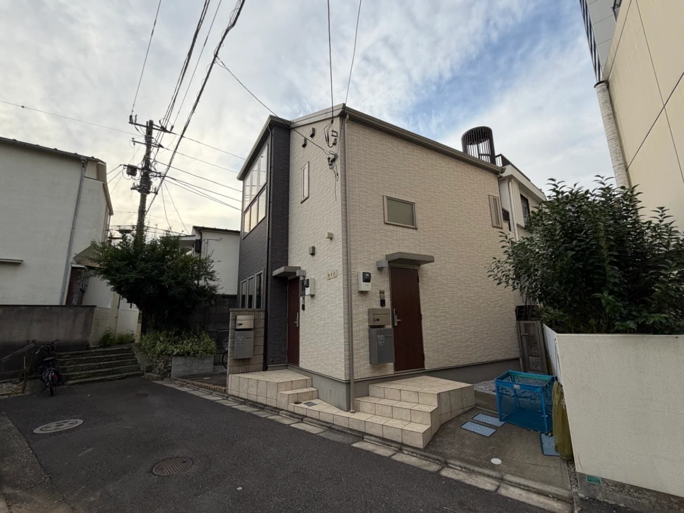 - 白とグレーの外壁が特徴の二階建て住宅、住宅街の角地に位置し、前には小さな階段と玄関ドア、周囲に樹木と配水管が見える