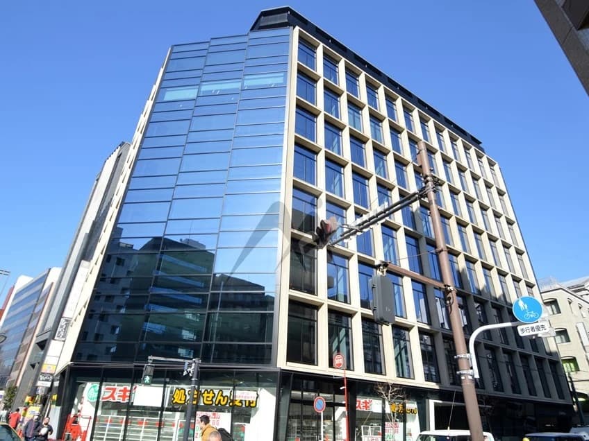 ガラス張りの四階建てオフィスビルの外観、青い空をバックに角部が強調されたモダンなファサードと街路景観、1階に店舗が並ぶ都市型ビル