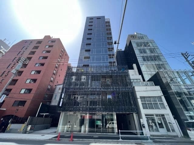 - ローカルな都市の高層ビル群が並ぶ street view の建物外観、正面に鉄骨のデザインゲートとガラス窓、晴天の青空と電線が上部を横切る風景