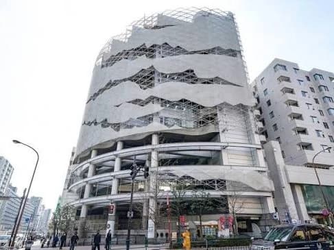 - 曲線的な白い外観の現代的ビル「ビル名風」, 階段状の波形デザインとガラスファサードが特徴的な建築物, 周囲の街並みと車両が写る都市景観の写真