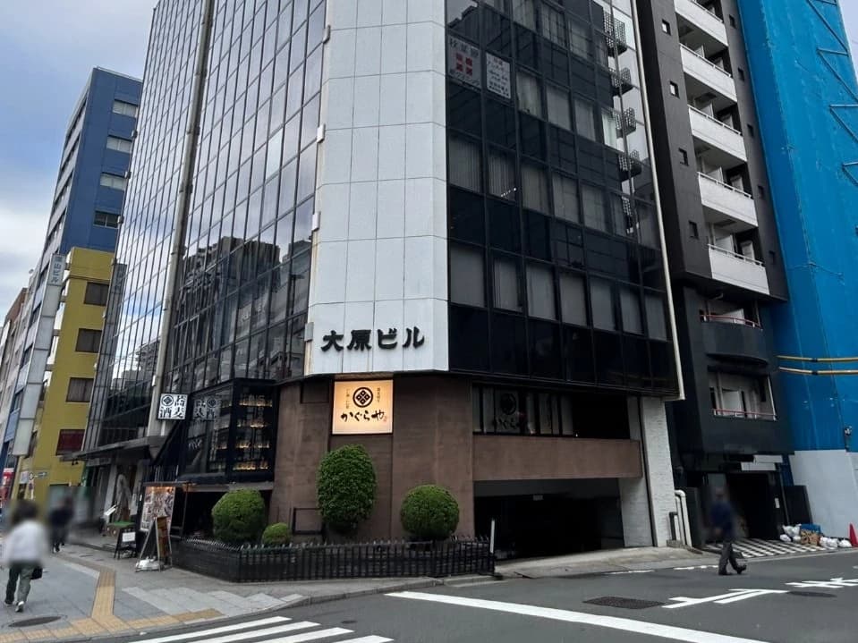 - 高層ビルが並ぶ新宿の街並み、ガラス張りの黒い外観と白色の看板が目を引くオフィスビル、路地沿いに緑の植栽と入口の暖簾がある風景