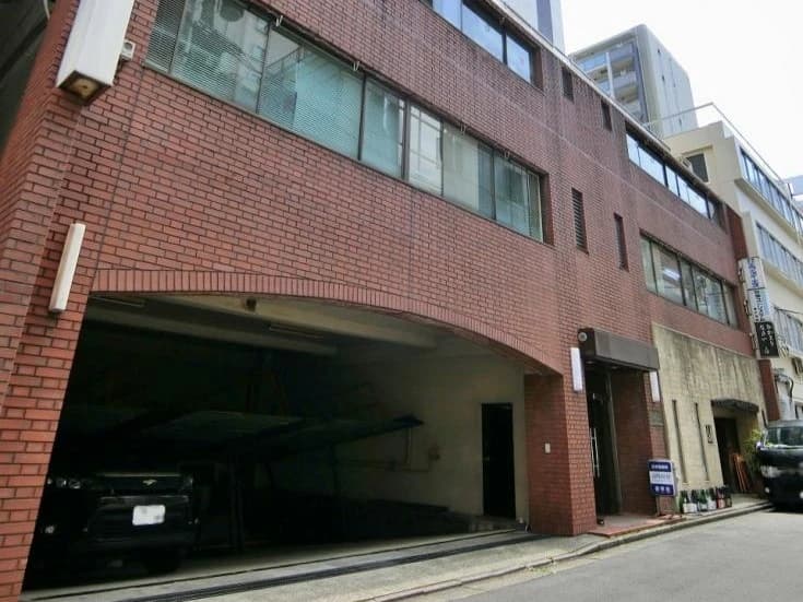 - 赤レンガ造りの三階建てビルの車庫付きファサード、路面側に駐車スペースと入口、小さな看板が並ぶ都会的な建物風景