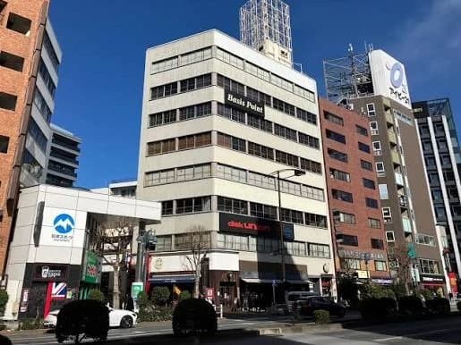 - 都市の商業ビル群が並ぶ街並み、中央の白いビルに「Bass Point」と表示、周囲に店鋪や看板、晴天の下で活気ある都市風景
