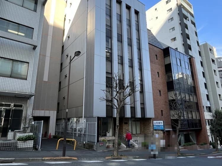- 都市の路地沿いに立つモダンなオフィスビル群、ガラス窓と縦長のシルバー外壁、左手の私道と歩道、手前に木と通行人が写るストリート風景