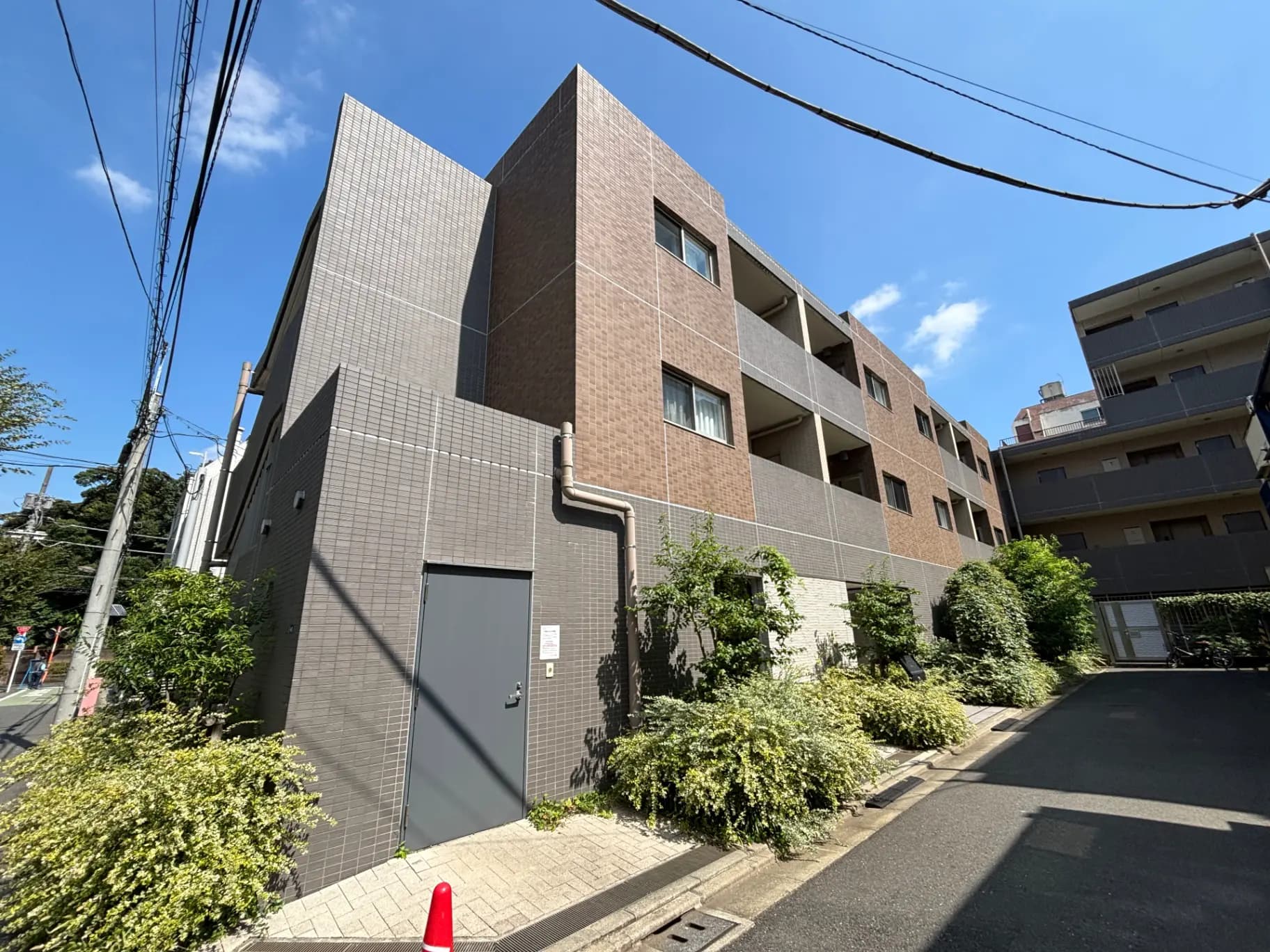 - モダンな外観のアパートメントビル、グレーと茶色のタイル張り、青空と緑豊かな植栽がある風景