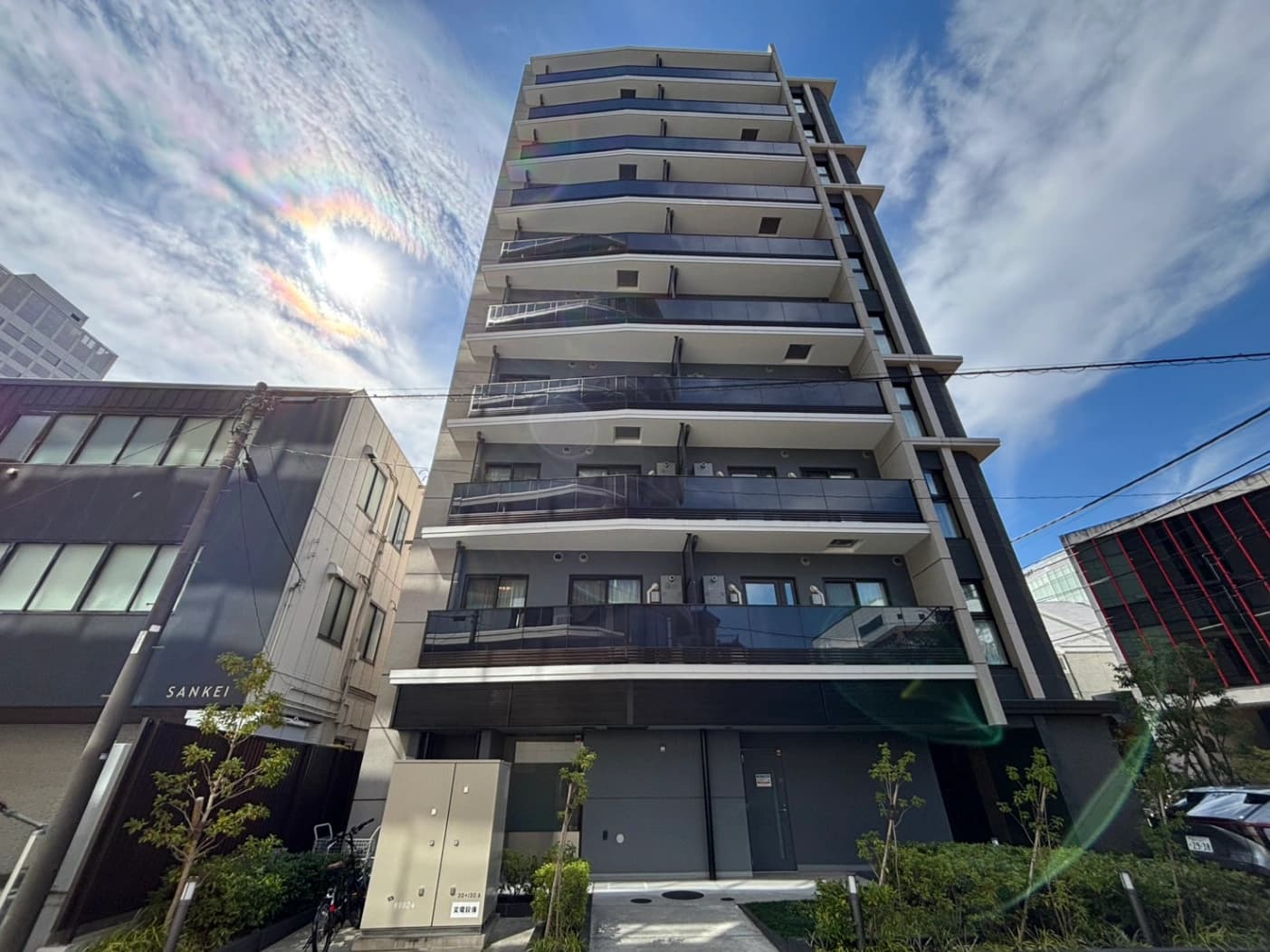 高層マンションの正面全景、複数階に渡るバルコニーとガラス手すり、周囲に低層の建物と緑豊かな植栽、晴天と青空を背景に強い日なたが差し込む都市景観