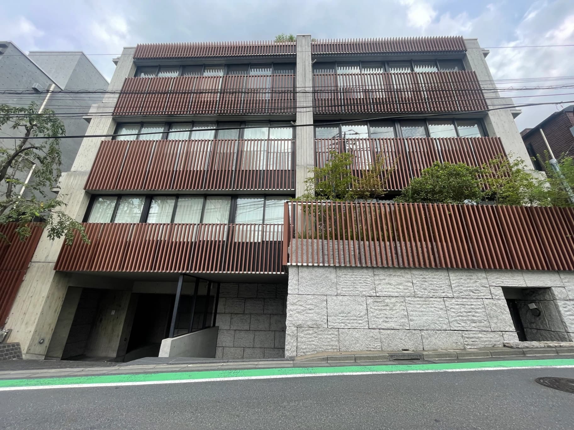 - コンクリートと木製ルーバーがモダンに組み合わさった三階建てのビル外観、縦長窓とグレーの石材壁、緑の樹木と電線が前景にある都市景観