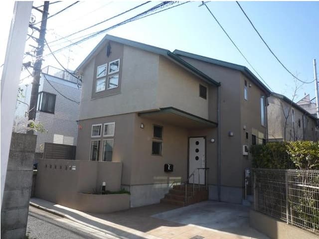 - 落ち着いたベージュの外観、階段のあるモダンな二階建て住宅、前面に小さな入口と窓が並ぶ suburban な家
- 住宅正面のショートアプローチ、コンクリートの駐車スペースと門扉、周囲に低い塀と金網のフェンスがある戸建て