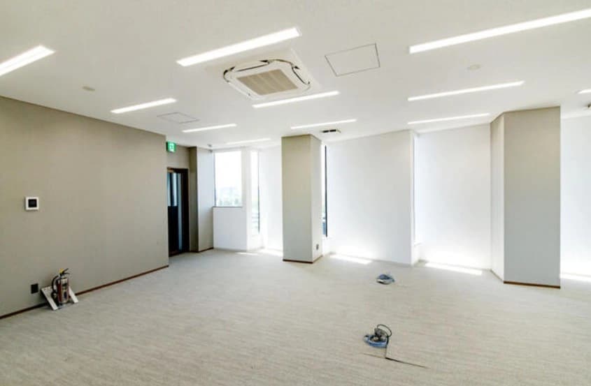 - オフィスのリノベーション途中の広い空間、白い壁と天井、列状の照明と窓から入る自然光、床には作業用コードと工具が散らばるシーン