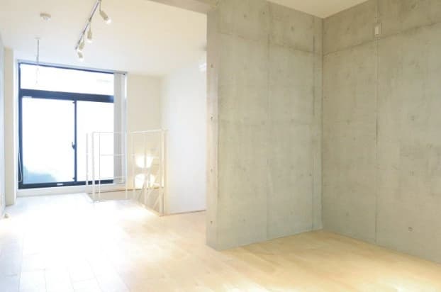 - コンクリート壁と木製フローリングのモダンな空間、開放的な窓辺と白い手すりの階段が見えるリビングエリア