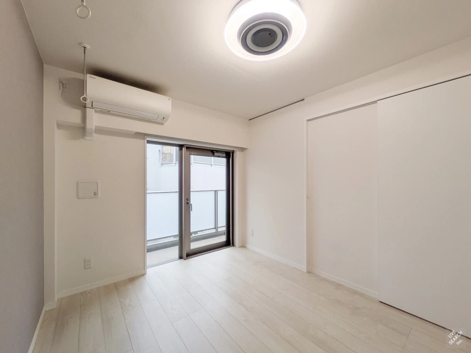 - 白を基調とした小さめの部屋、壁掛けエアコンと大きな窓付きのリビング、床は淡い木目調、スライドドアの収納スペースと間接照明の設置