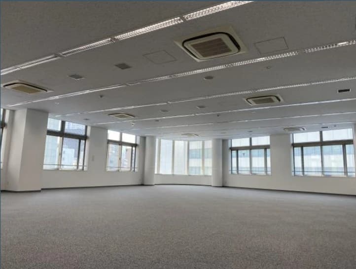 - 広々としたオフィスフロア、白い壁と大きな窓から差し込む自然光、ビル屋内の無人空間